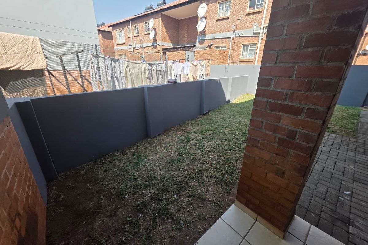 A private rear garden area featuring a paved walkway, a small lawn, and a brick pillar supporting a covered patio section.
