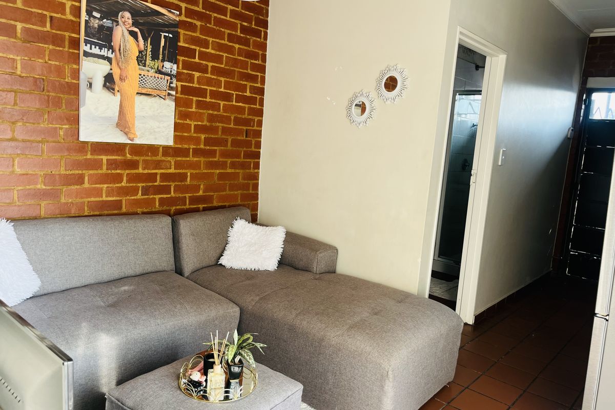 A living room featuring an exposed brick feature wall and tiled flooring, with an adjacent doorway leading to a bathroom area.