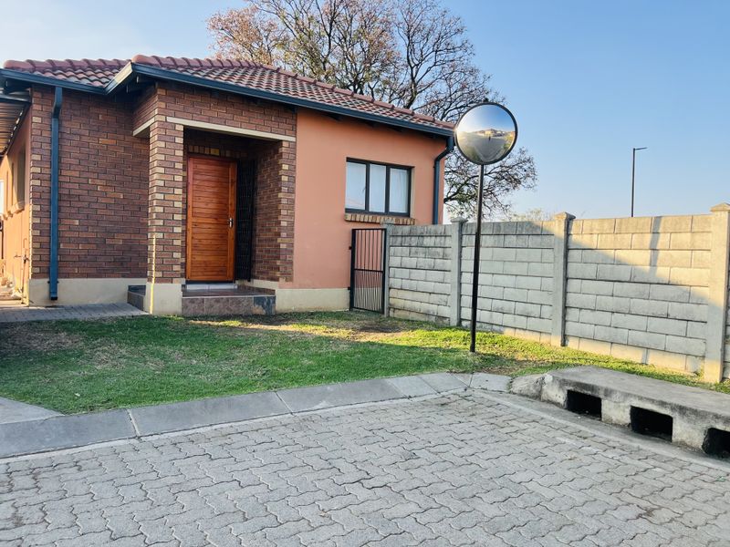 Front view of a brick townhouse featuring a wooden entrance door, paved driveway, and concrete boundary wall with a safety mirror.