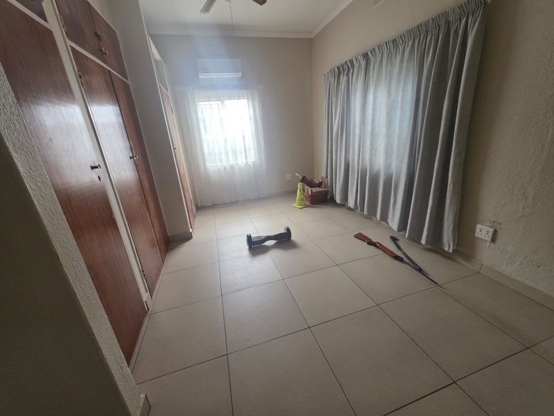 A spacious bedroom featuring tiled flooring, built-in wooden cupboards, a ceiling fan, and a wall-mounted air conditioning unit.