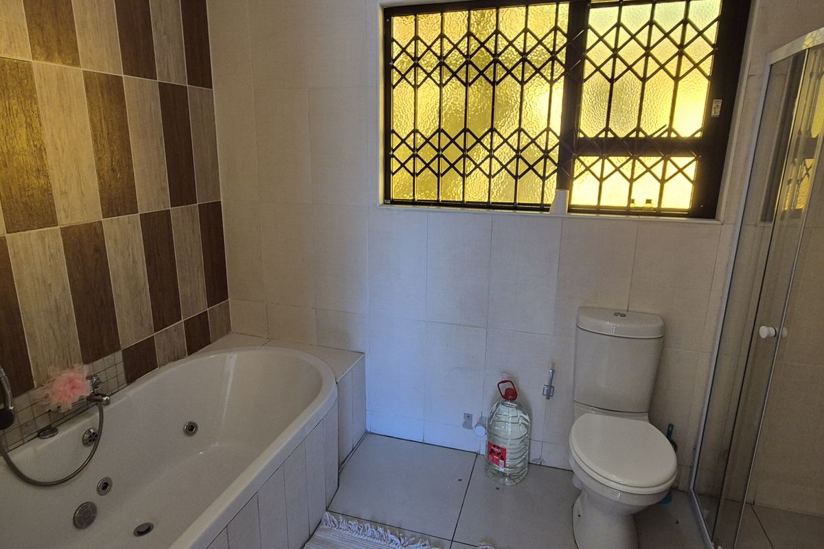 A functional bathroom featuring a built-in jetted bathtub, separate shower enclosure, toilet, and tiled finishes with a security-gated window.