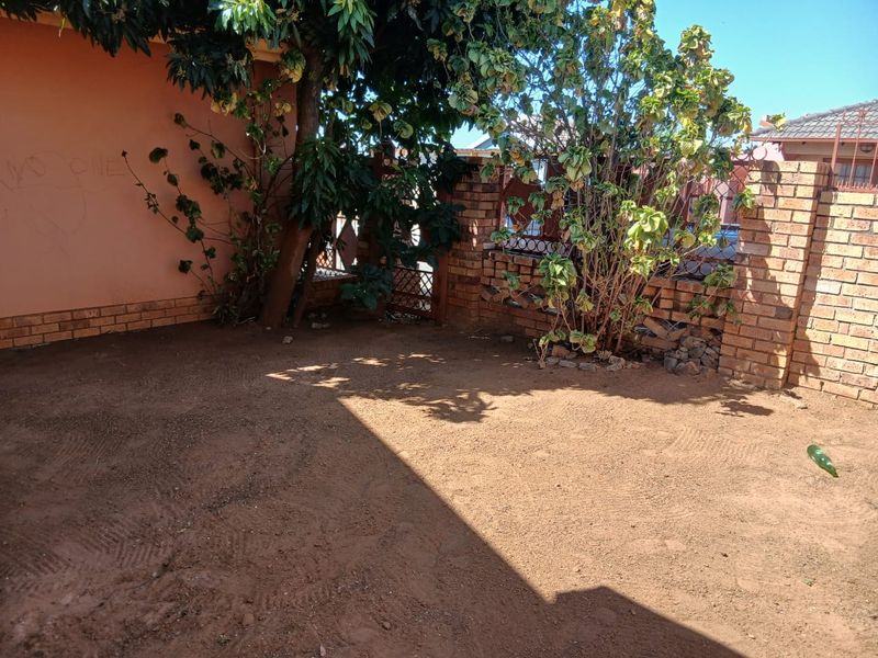 A rear outdoor area featuring a brick boundary wall, metal gate, and unlandscaped soil ground, requiring maintenance and landscaping improvements.
