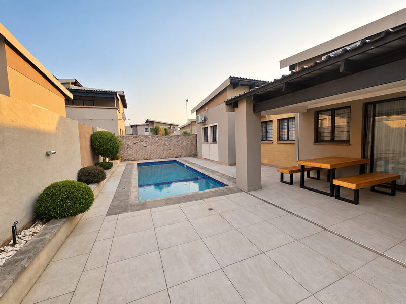 A well-maintained outdoor entertainment area featuring a swimming pool, tiled patio, and covered seating space, enclosed by a brick boundary wall.