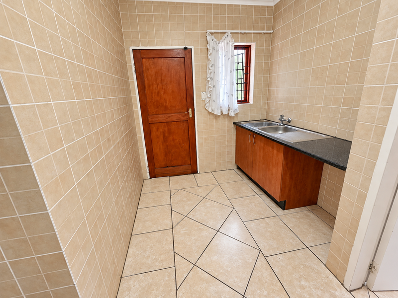 A functional scullery featuring full-height wall tiling, tiled flooring, built-in cabinetry with a stainless steel sink, and a solid timber door.