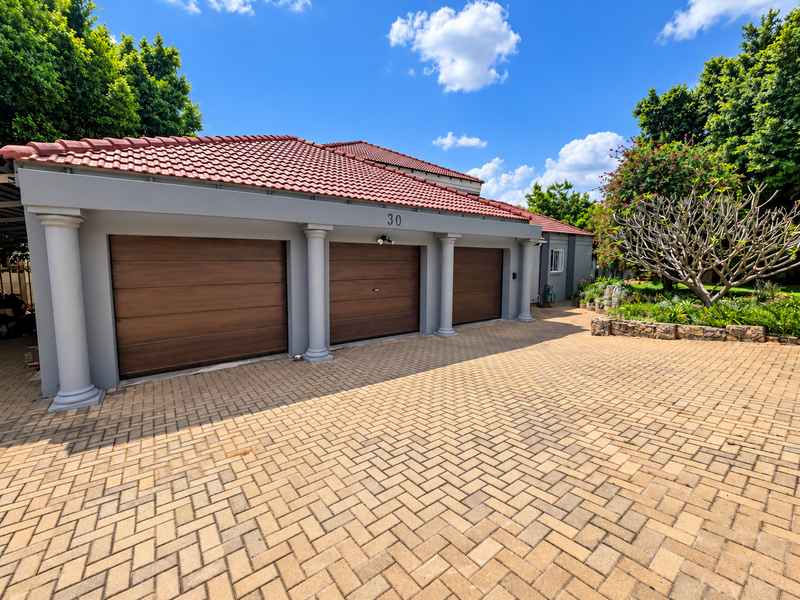 A well-maintained triple garage with modern wooden-finish doors, decorative columns, and a spacious paved driveway set in a landscaped garden.