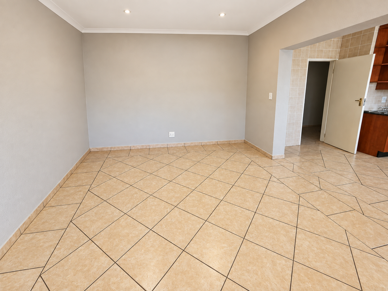 A bright, empty living area featuring neutral walls, tiled flooring laid on a diagonal, and recessed downlighting, with an adjacent kitchen doorway.