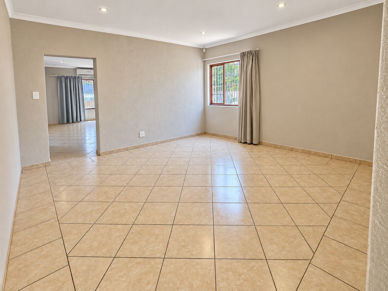 A spacious, empty living room featuring neutral tiled flooring, recessed downlighting, and a window with security bars, offering a clean and versatile living space.