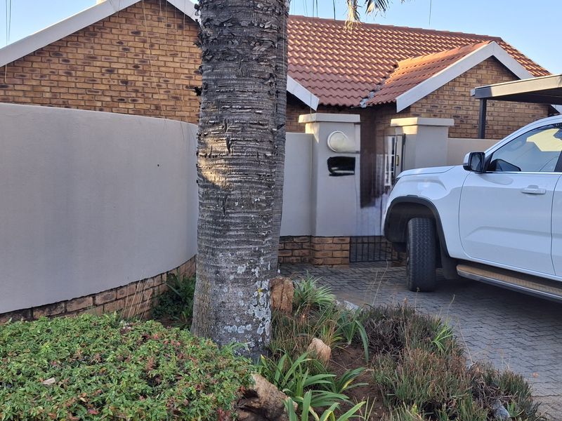 Front view of a brick house featuring a tiled roof, paved driveway, and a carport, enclosed by a boundary wall.