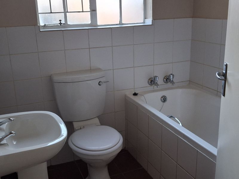 A functional bathroom featuring a pedestal basin, toilet, and bathtub with white wall tiling and a window with security bars.