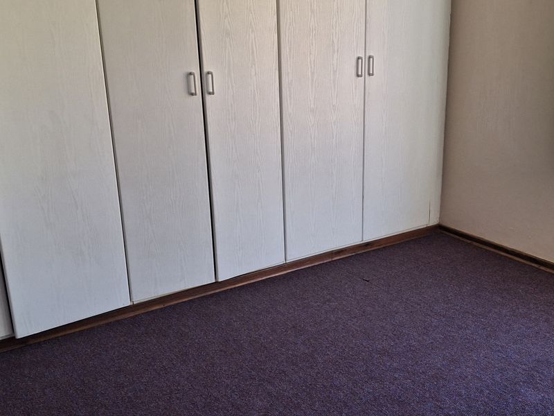 A vacant bedroom featuring built-in cupboards, carpeted flooring, and a window fitted with security burglar bars.