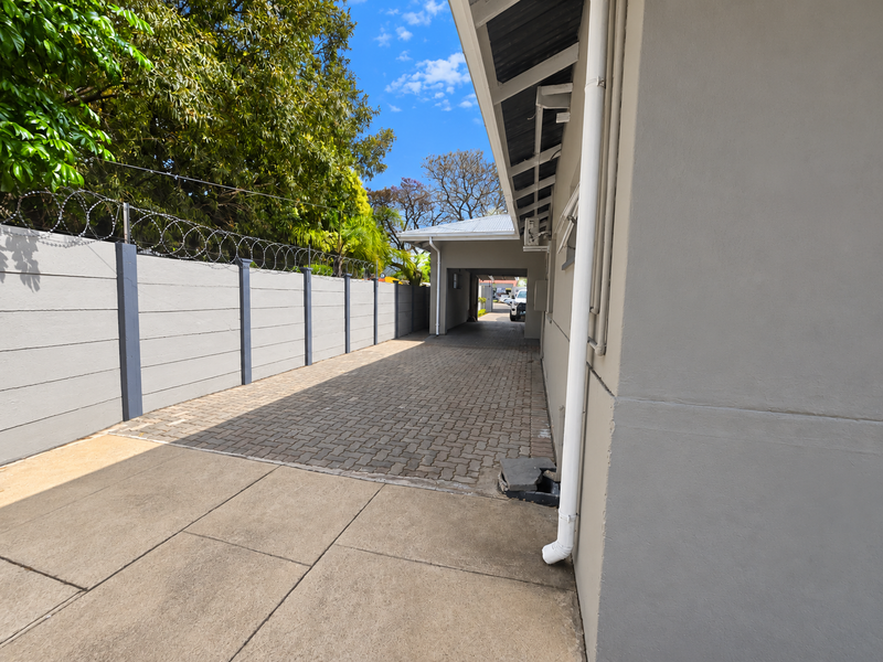 A paved side driveway leading to a covered carport, featuring a secure boundary wall with razor wire and functional drainage infrastructure.