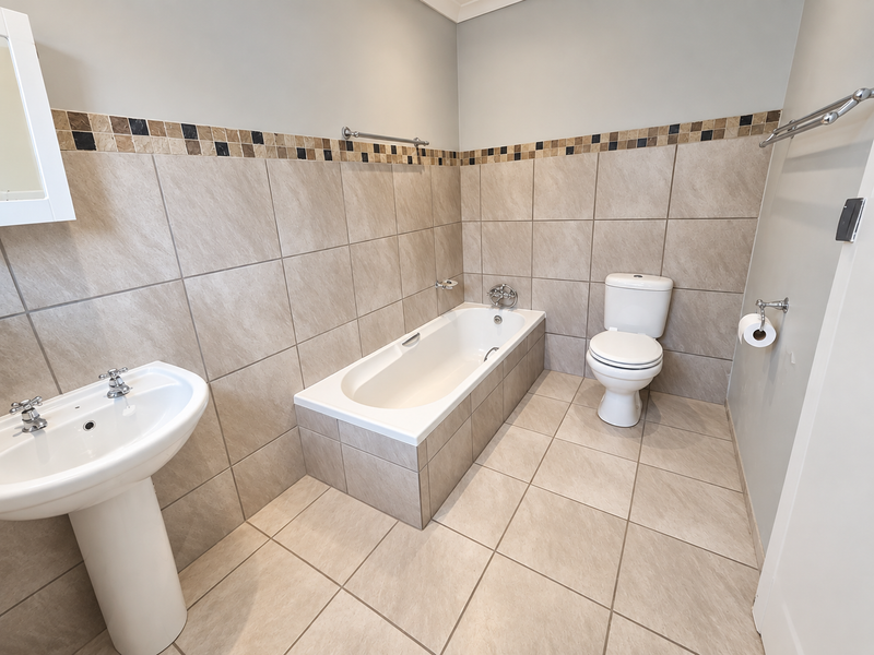 A clean, neutral-toned bathroom featuring a pedestal basin, a built-in bathtub, and a toilet, finished with tiled walls and a decorative mosaic border.