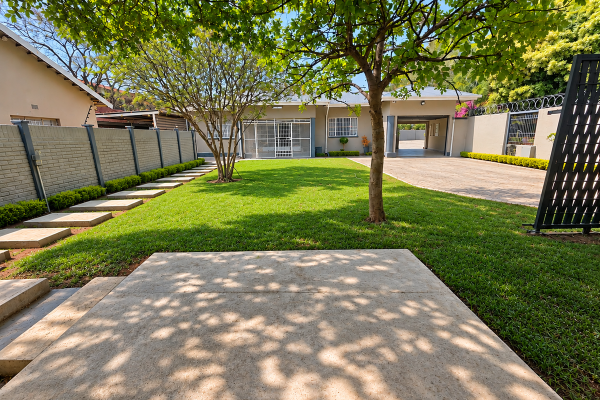 A well-maintained rear garden featuring a lush lawn, paved driveway, concrete patio area, and secure boundary walls with integrated security features.