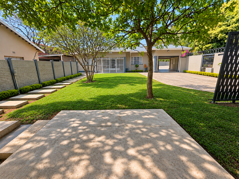 A well-maintained rear garden featuring a lush lawn, paved driveway, concrete patio area, and secure boundary walls with integrated security features.