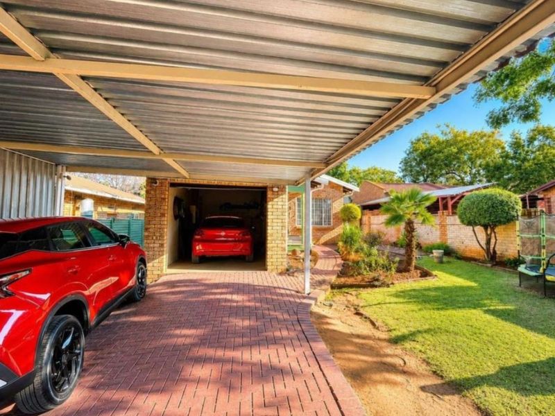 A paved driveway leading to a brick garage with a corrugated metal carport, set alongside a landscaped garden and brick boundary wall.