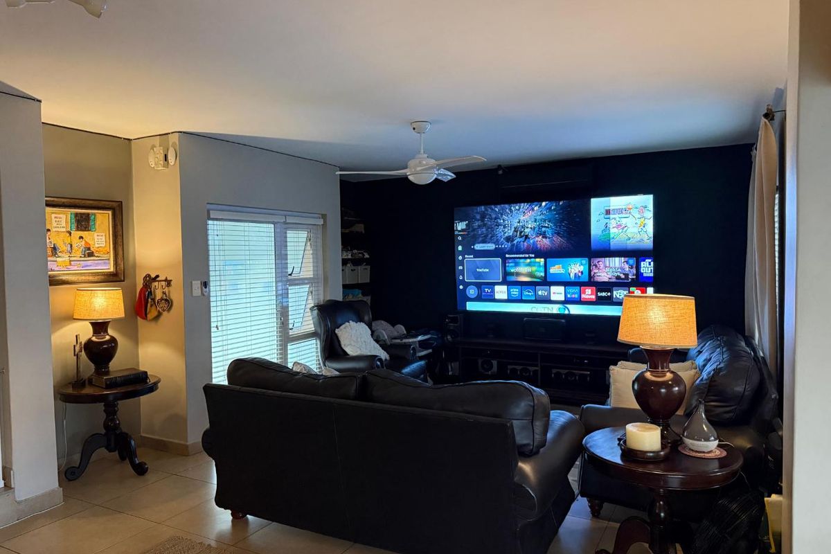 A comfortable living room featuring tiled flooring, a ceiling fan, and an air conditioning unit, with a large wall-mounted television as the focal point.