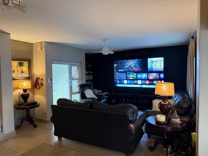 A comfortable living room featuring tiled flooring, a ceiling fan, and an air conditioning unit, with a large wall-mounted television as the focal point.