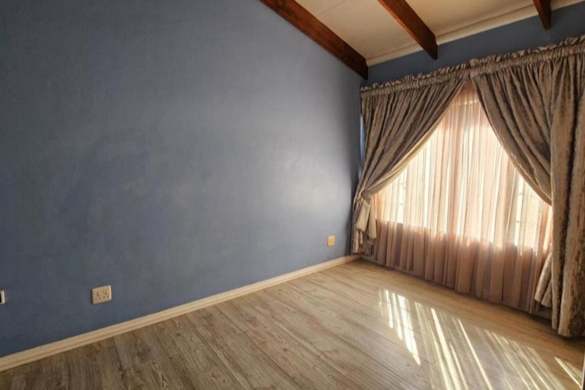 A bright, empty bedroom featuring laminate flooring, exposed timber ceiling beams, and a large window with curtains.
