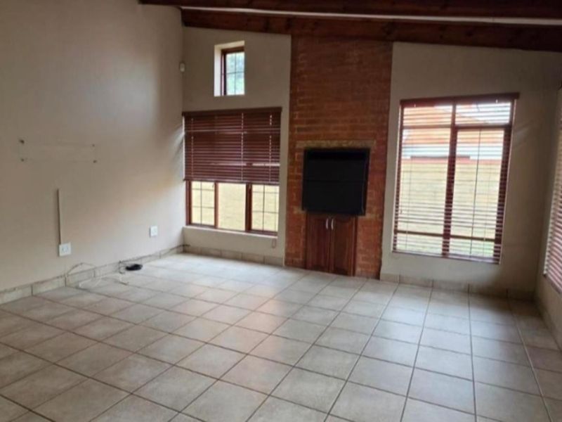 Spacious living room featuring exposed timber ceiling beams, a brick fireplace, tiled flooring, and large windows with venetian blinds.