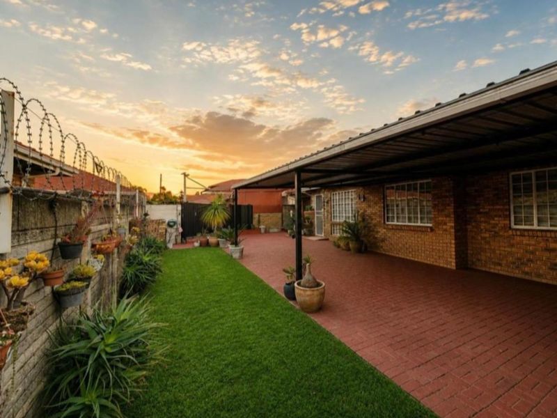 A well-maintained rear exterior area featuring a paved carport, a neat lawn, and a secure brick boundary wall with razor wire.