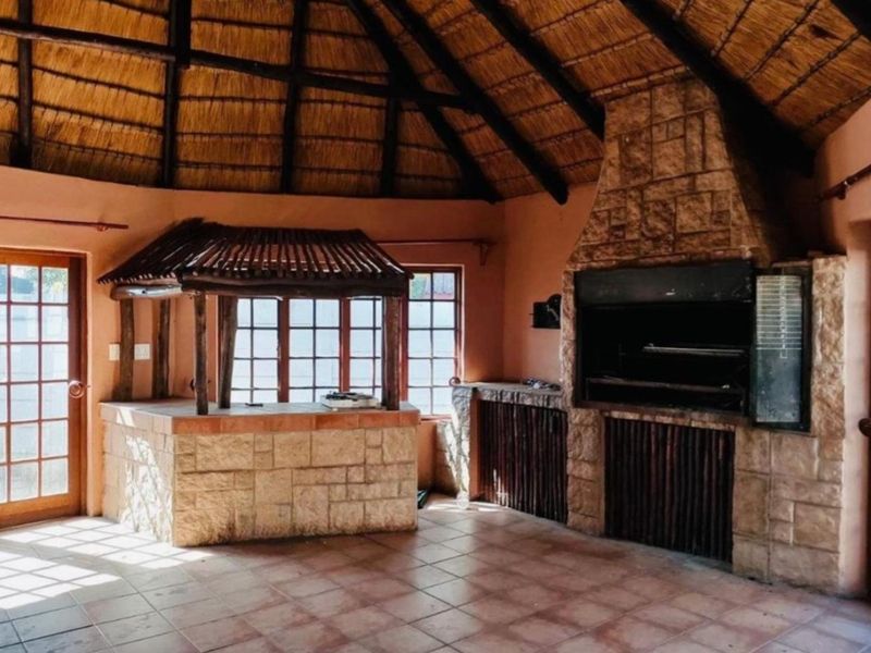 Spacious covered entertainment area featuring a traditional thatched roof, built-in stone braai, and a stone-clad bar counter with tiled flooring.
