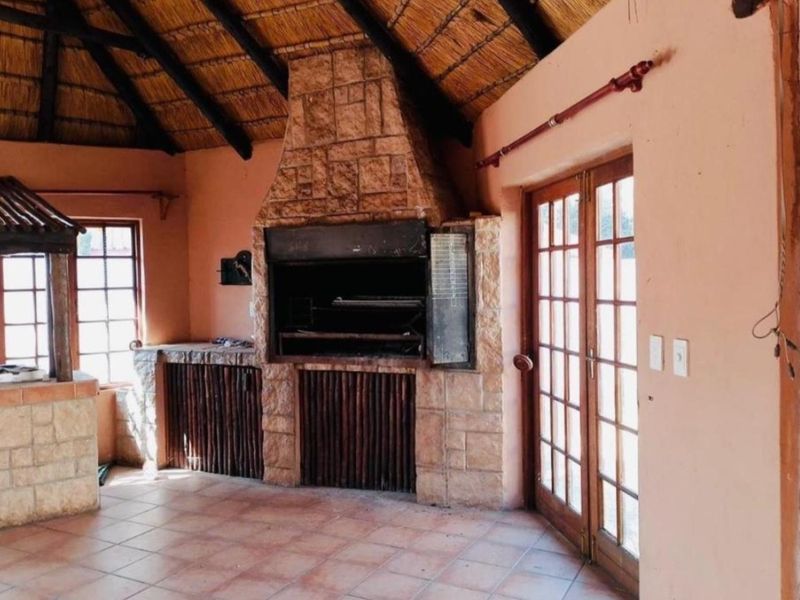Covered patio area featuring a large built-in stone braai, thatched roof with exposed beams, and tiled flooring, offering a rustic outdoor entertainment space.