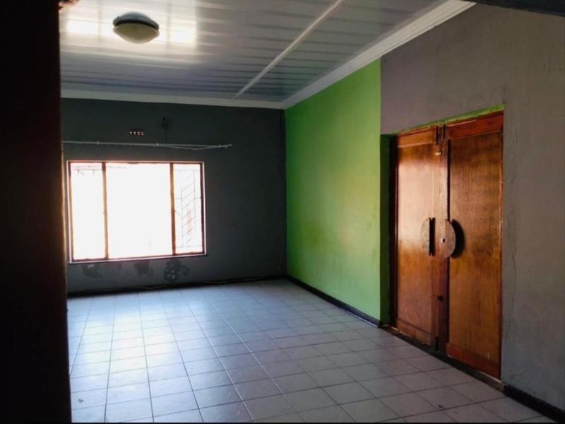 An empty living room featuring tiled flooring, double wooden doors, and a window with security bars, offering a blank canvas for renovation.
