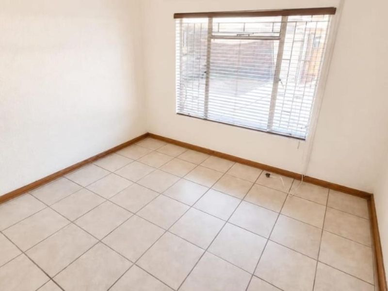 A bright, empty bedroom featuring neutral tiled flooring, clean walls, and a window fitted with security bars and venetian blinds.