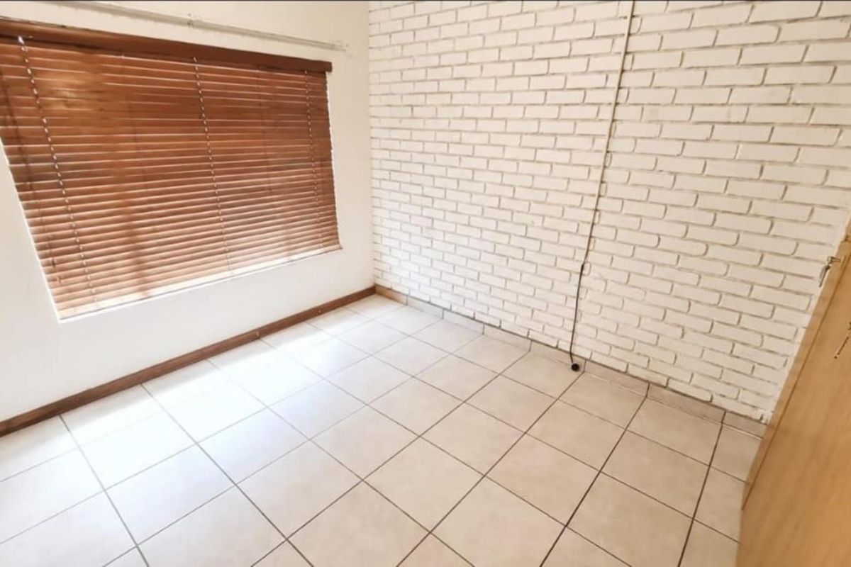 A bright, empty bedroom featuring tiled flooring, a white painted brick feature wall, and wooden venetian blinds.