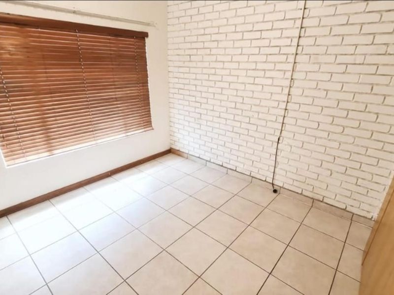 A bright, empty bedroom featuring tiled flooring, a white painted brick feature wall, and wooden venetian blinds.