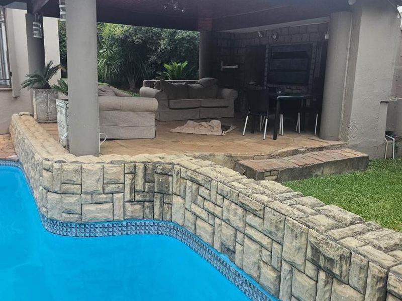 Outdoor entertainment area featuring a covered patio with a built-in braai, overlooking a swimming pool with a decorative stone-clad retaining wall.