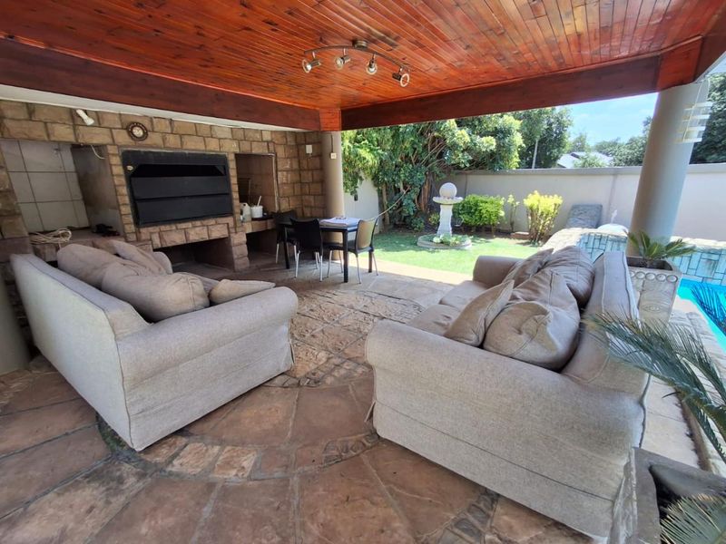 Covered outdoor patio featuring a built-in braai, stone cladding, and timber ceiling, overlooking a private garden and swimming pool area.