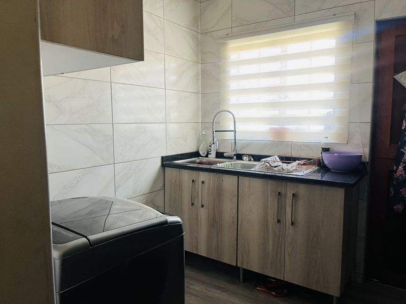 A functional laundry area featuring modern wood-finish cabinetry, granite countertops, a stainless steel sink with a gooseneck mixer, and elegant marble-look wall tiling.