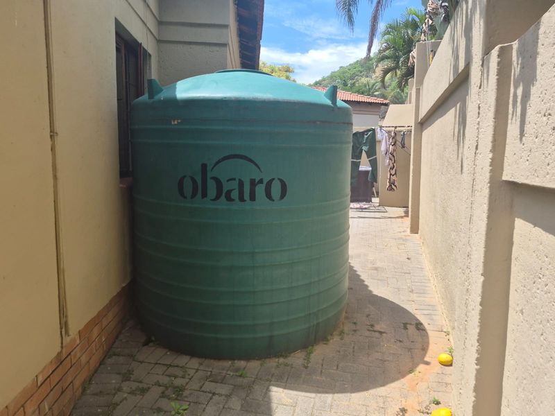 A side exterior area featuring a large green water storage tank situated on a paved walkway alongside the property boundary wall.