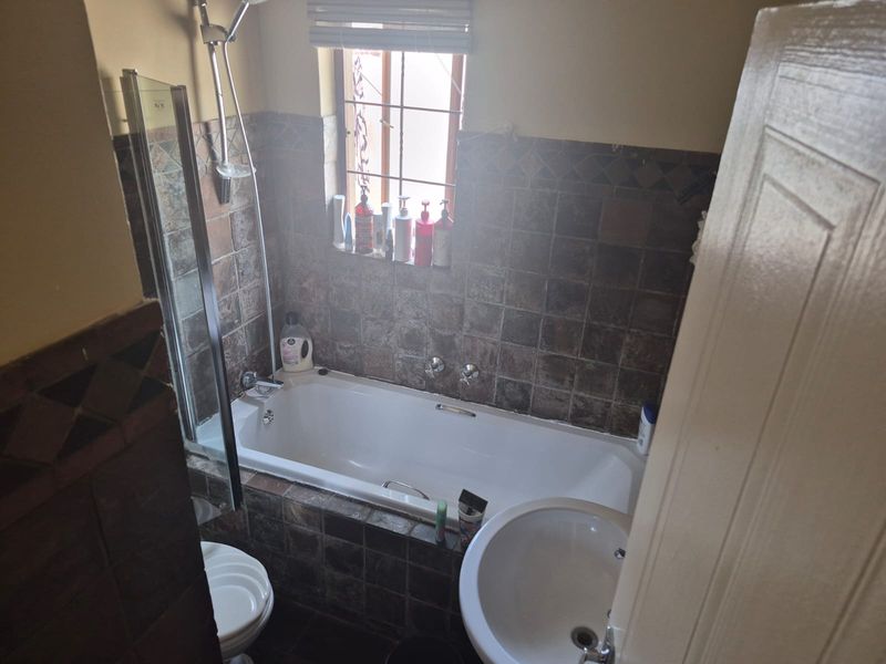 A compact bathroom featuring a built-in bathtub with shower attachment, pedestal basin, and toilet, finished with dark decorative wall tiling.