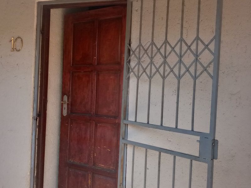 Entrance area of the flat featuring a timber door and a metal security gate, with visible wear on the door and exterior wall finish.