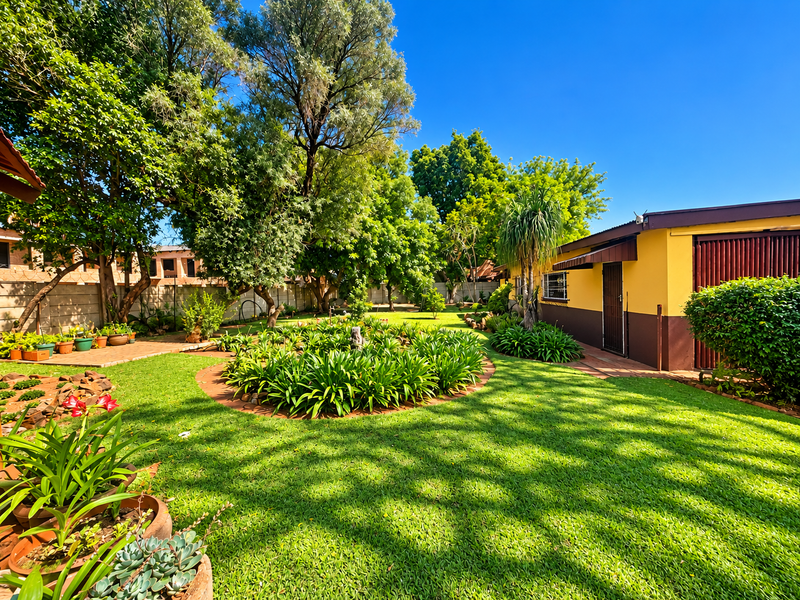 A well-maintained backyard garden featuring lush green lawn, mature trees, and established garden beds alongside the rear exterior of the house.