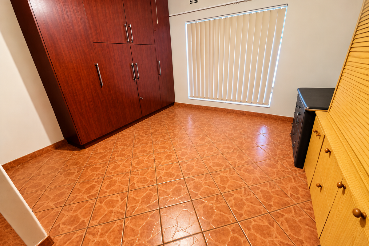 A bedroom featuring dark wood built-in cupboards, terracotta-style tiled flooring, and a window fitted with vertical blinds.