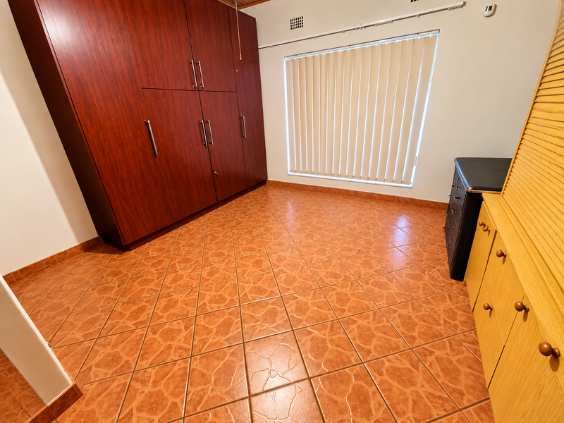 A bedroom featuring dark wood built-in cupboards, terracotta-style tiled flooring, and a window fitted with vertical blinds.