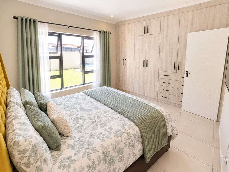 A well-maintained bedroom featuring neutral tiled flooring, ample built-in cupboard storage, and large aluminium windows overlooking the garden.