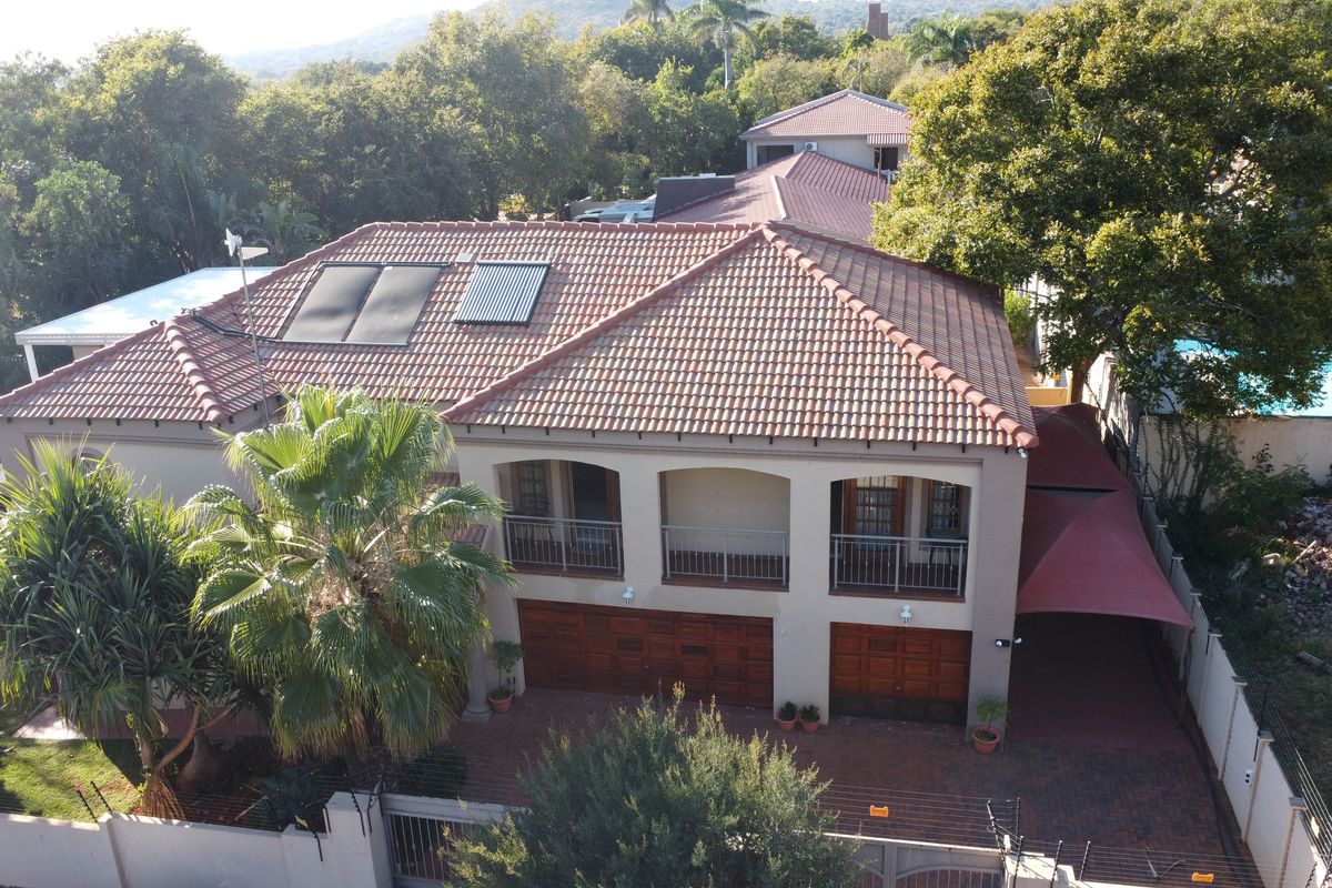 Aerial view of a residential property featuring a tiled roof with solar panels, double garage, balcony, and paved driveway within a landscaped setting.