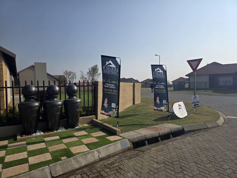 Street view of the townhouse complex entrance featuring a paved driveway, boundary wall, and decorative water feature.