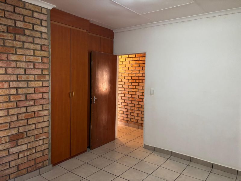 A vacant bedroom featuring tiled flooring, built-in wooden cupboards, and an exposed brick feature wall, leading into an adjacent brick-walled space.
