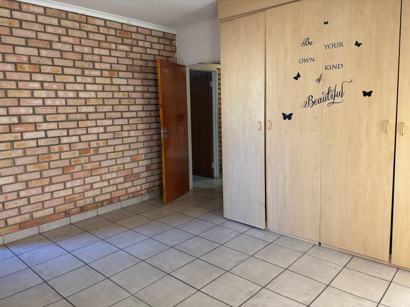 A spacious bedroom featuring a distinctive exposed brick wall, neutral tiled flooring, and ample built-in cupboard storage.