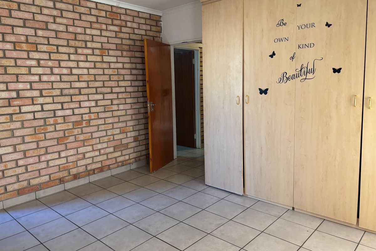 A spacious bedroom featuring a distinctive exposed brick wall, neutral tiled flooring, and ample built-in cupboard storage.