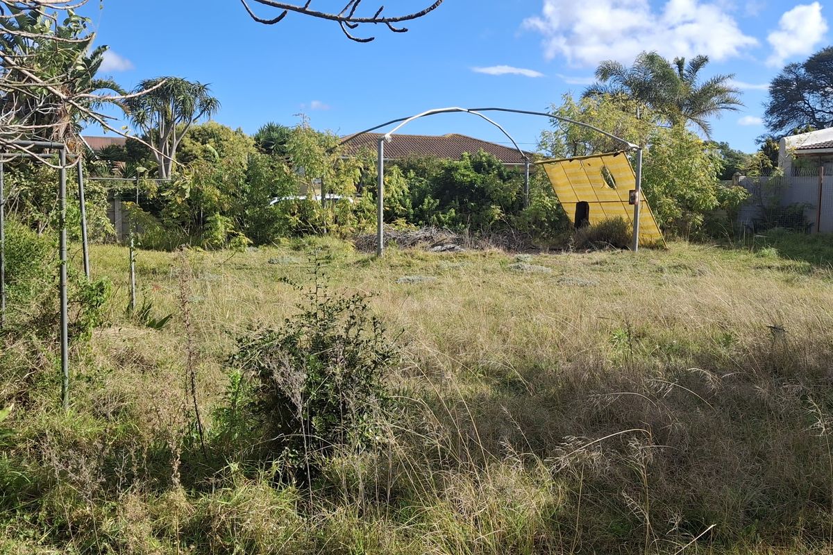 A vacant residential plot featuring overgrown grass, mature trees, perimeter fencing, and a damaged metal frame structure.