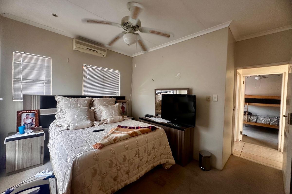 A bedroom featuring a ceiling fan, wall-mounted air conditioning unit, and venetian blinds, with an adjacent room visible through the doorway.