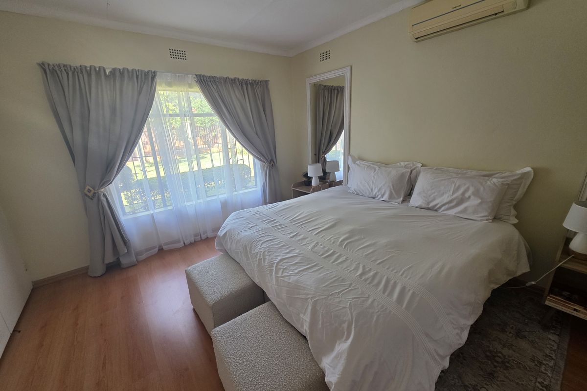 A well-lit bedroom featuring laminate flooring, a wall-mounted air conditioning unit, and a large window with curtains overlooking the garden.