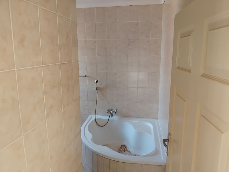 A compact bathroom featuring a corner bathtub with wall-mounted handheld shower fixtures and neutral ceramic wall tiling.
