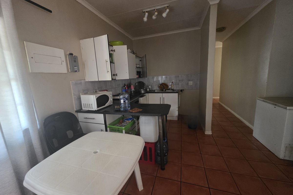 A compact kitchen area featuring basic cabinetry, tiled splashbacks, and terracotta floor tiles, leading into a hallway.